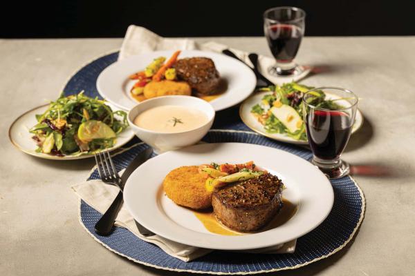 Two place settings with a fillet mignon, risotto cakes, lobster bisque, red wine, and side salads