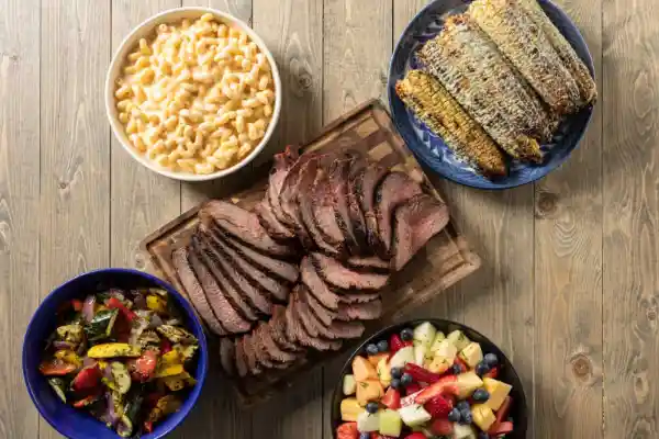 Sliced tri-tip on cutting board next to grilled corn, macaroni and cheese, a fruit salad and grilled vegetables 