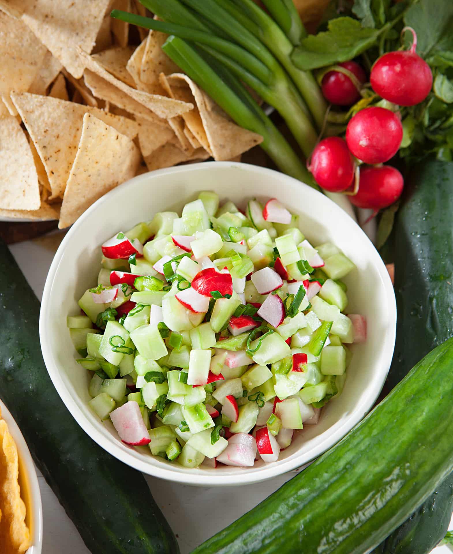 Cucumber salsa in a white bowl surrounded by tortilla chips and ingredients
